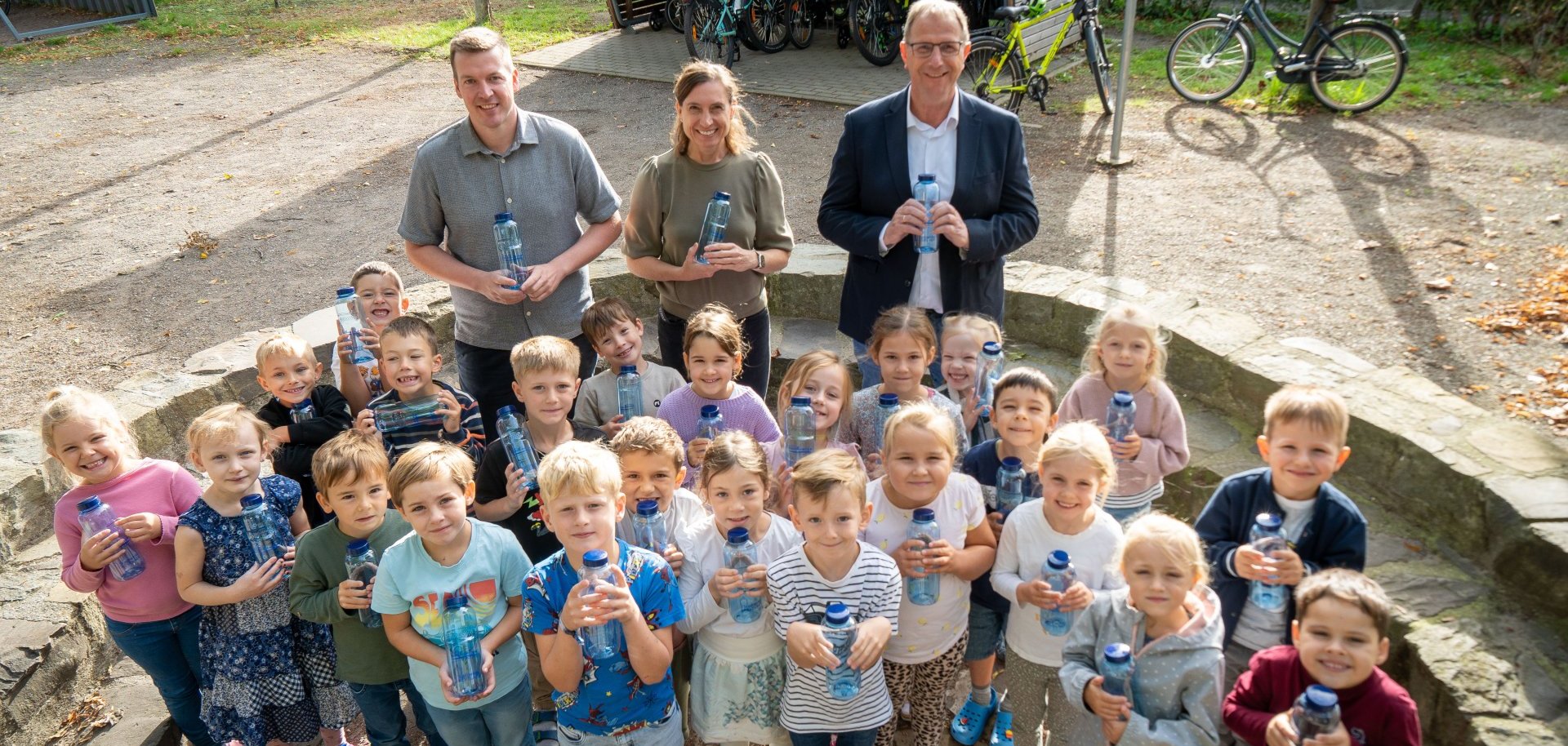 First-graders at St. Franziskus Elementary School in Twisteden are standing in the schoolyard with their new blue water bottles. Behind them are Hendrik Kruß, Principal Tanja Kocken, and Wolfgang Toonen.