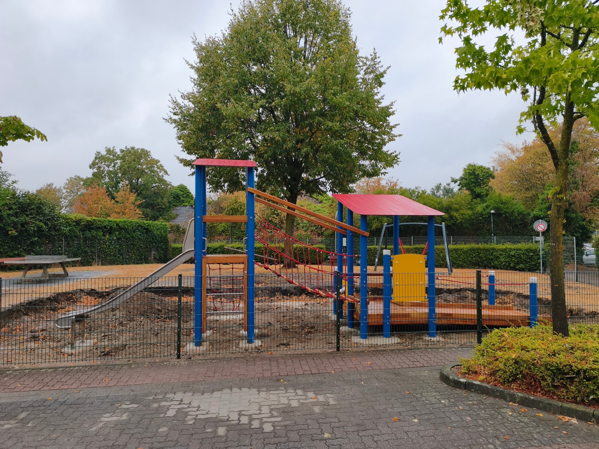 New blue, red, and yellow playground equipment at the playground on Theodor-Heuss-Ring, featuring a slide, climbing structures, a rope suspension bridge, and a wheelchair-accessible walkway.