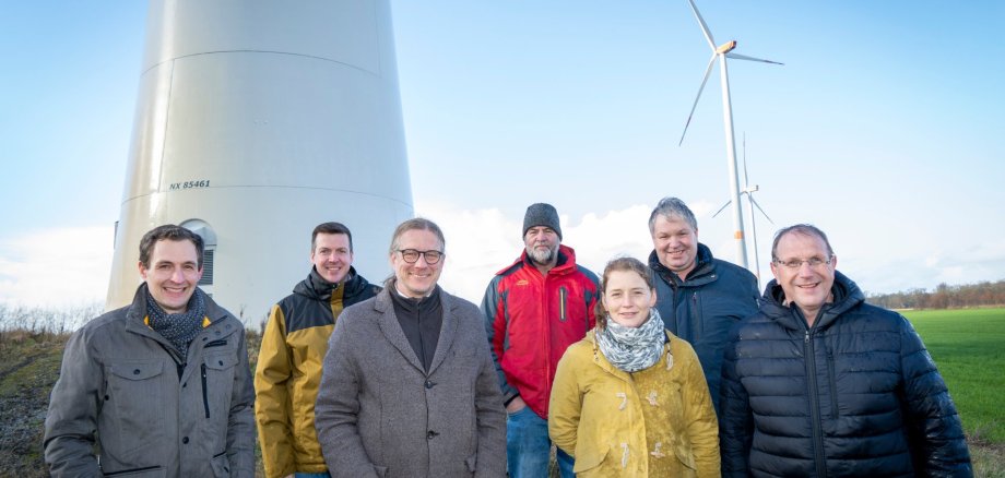 Zeven vertegenwoordigers van de fondsinvesteerders staan voor een windturbine in Kevelaer. Op de achtergrond zijn andere windturbines en een open landschap te zien. De foto symboliseert de promotie van liefdadigheidsprojecten met de opbrengst van lokale windenergie.