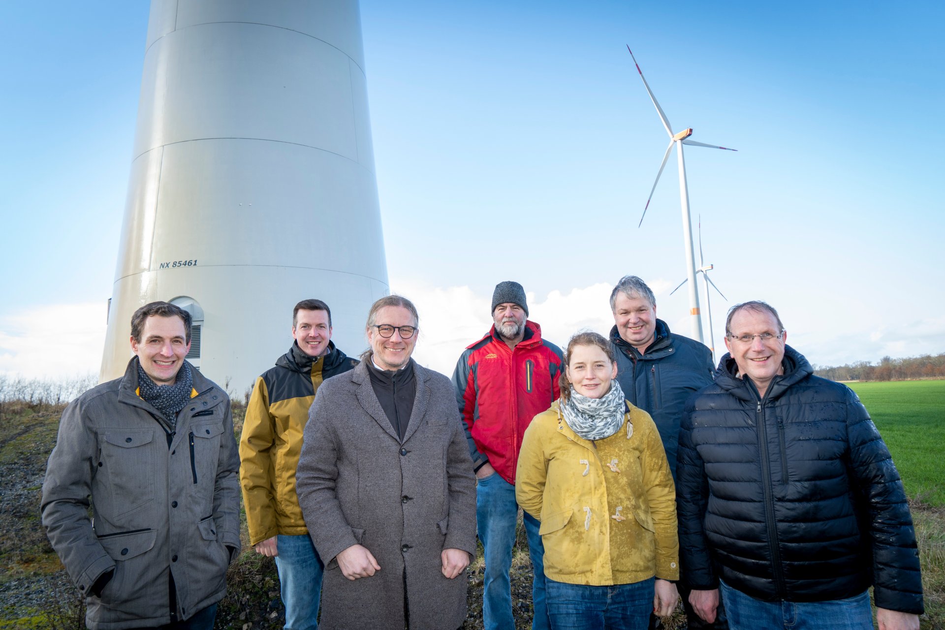 Zeven vertegenwoordigers van de fondsinvesteerders staan voor een windturbine in Kevelaer. Op de achtergrond zijn andere windturbines en een open landschap te zien. De foto symboliseert de promotie van liefdadigheidsprojecten met de opbrengst van lokale windenergie.