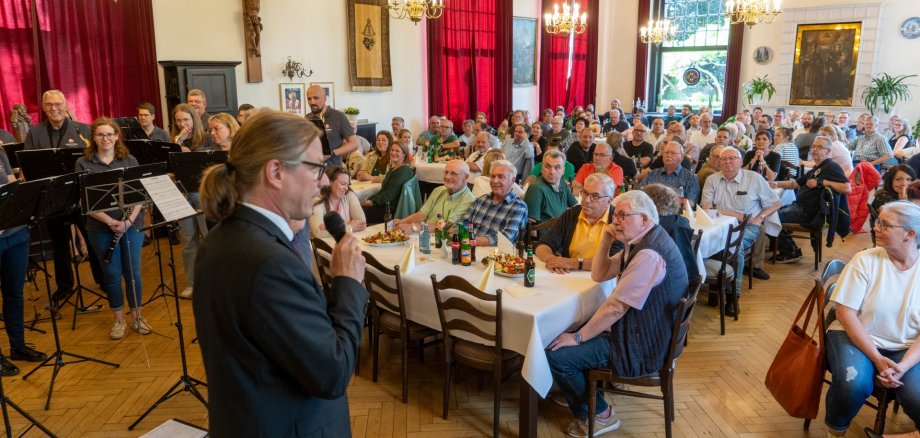 Een man spreekt tot mensen die aan tafels in een hal zitten.